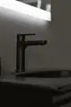 Schwarze Waschtischarmatur von Ideal Standard, minimalistische Form, montiert auf einer dunklen Oberfläche, neben Waschbecken und beleuchtetem Spiegel.